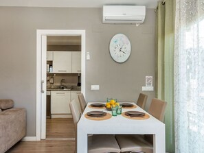 Une salle à manger avec une table blanche dressée pour quatre personnes, une horloge au mur et une cuisine en arrière-plan.