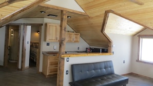 Une chambre aménagée dans les combles, avec un plafond en pente, des poutres apparentes, un banc en cuir noir, un coin cuisine doté de meubles en bois et une fenêtre.