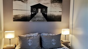 Een slaapkamer met een bed, twee bedlampjes, een kunstwerk aan de muur van een huis op een pier, en een plafondlamp.