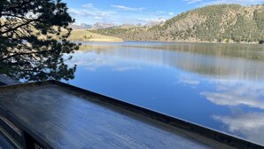 A wooden deck with "WELCOME TO HOLTER" engraved, overlooking a calm lake with mountains in the background. - Holter Lake Water Front Cabin (Wolf Creek)