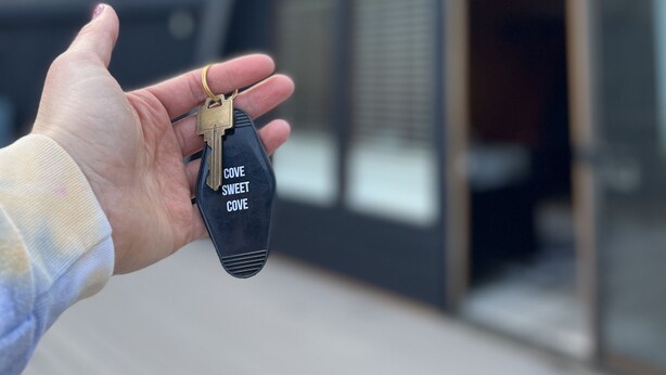 A hand holding a key with the inscription "Cove Sweet Cove" in front of a modern house with large windows and a sloped roof. - Holter Lake Water Front Cabin (Wolf Creek)
