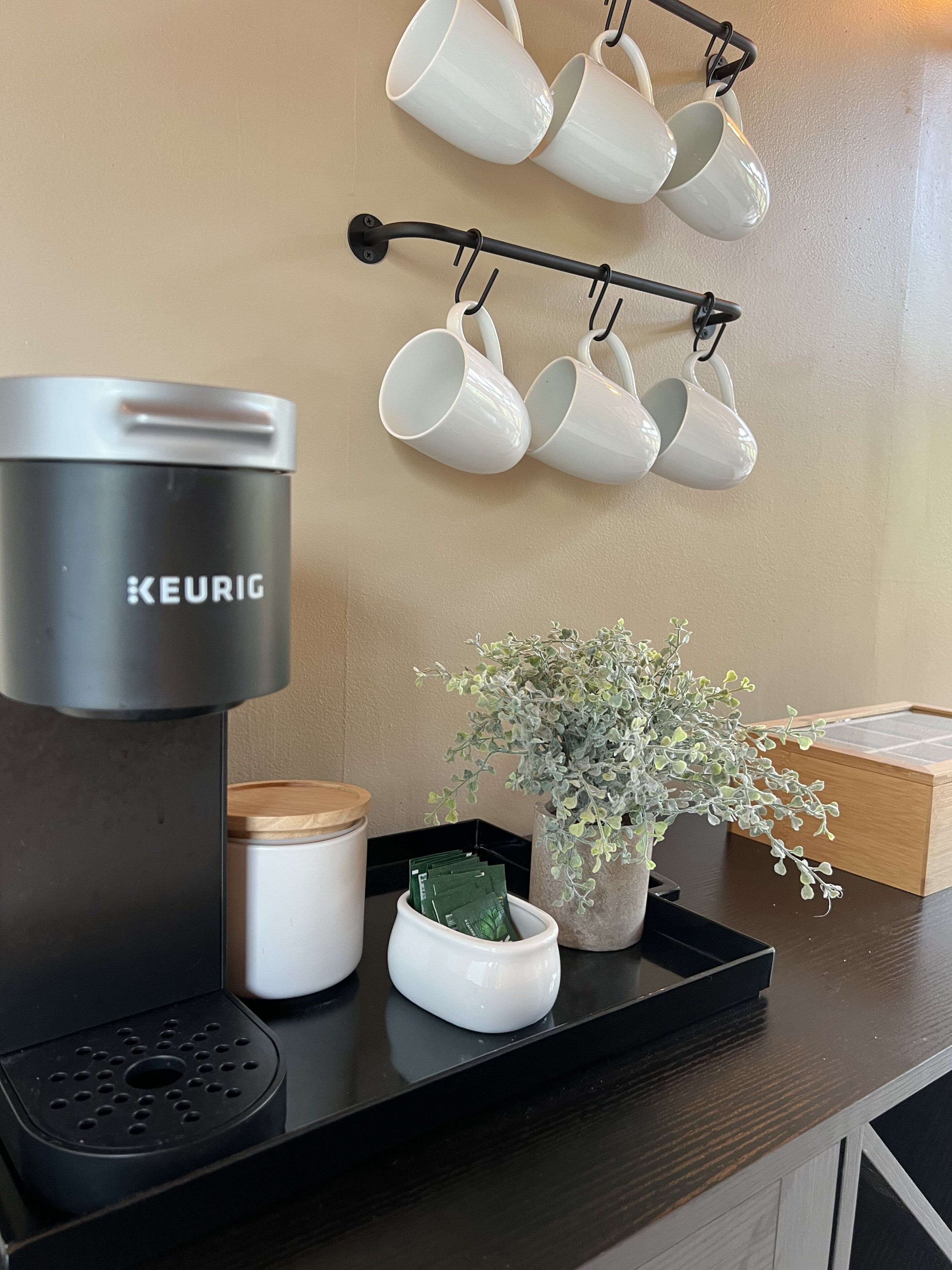 A kitchen counter with a Keurig coffee maker, a white mug rack, a potted plant, and a wooden box.