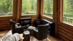 A log cabin interior with a wooden ceiling, large windows, a ceiling fan, and a seating area with a bookshelf and ottoman. - Cozy Log Cabin Near Indiana Dunes & Lake Michigan! (Portage)