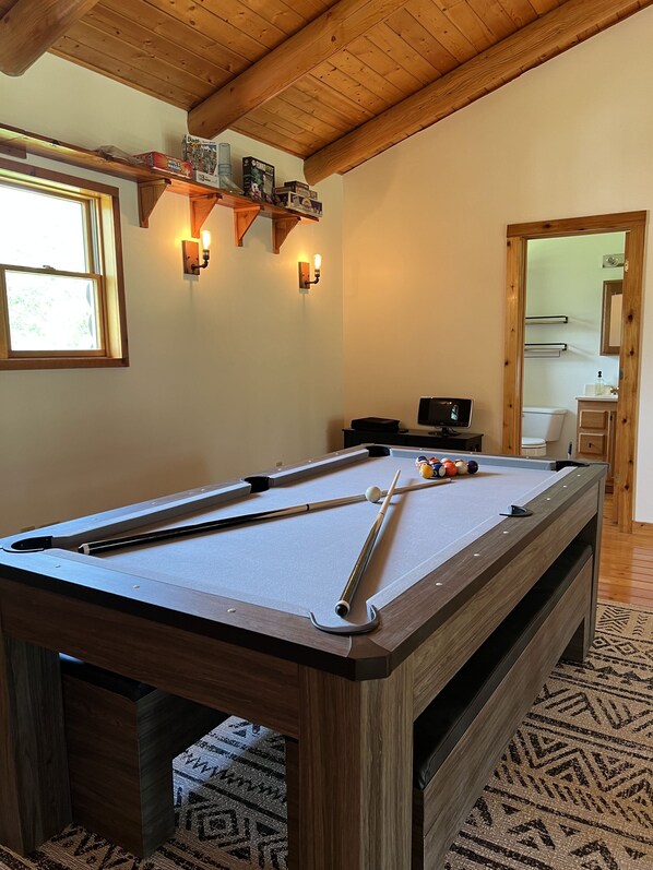 A pool table with cues and balls, wooden beams, a window, and a mirror in a room with a patterned rug.