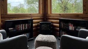 A cozy cabin interior with wooden walls, two large windows offering a view of trees and sky, a small round table, and two chairs. - Cozy Log Cabin Near Indiana Dunes & Lake Michigan! (Portage)