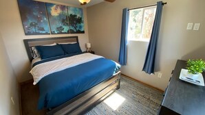 Un dormitorio con cama, ventilador de techo, pintura de un cielo nocturno, ventana con cortinas y una mesita con una planta.