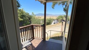 Vista desde la puerta de una habitación a una terraza exterior con baranda de madera, una silla y cielo despejado con montañas a lo lejos.