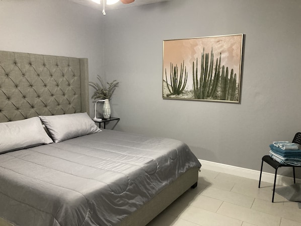 A bedroom with a tufted headboard, a bed with grey bedding, a bedside table with a lamp and a vase, and a framed picture of cacti on the wall.