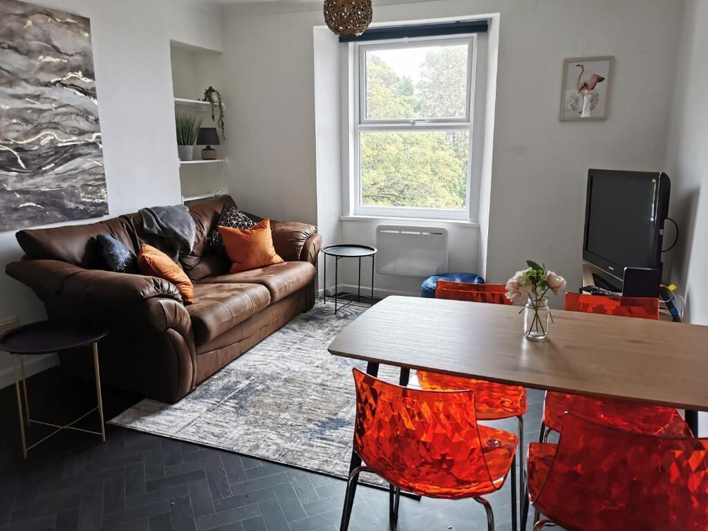 Ein modernes Wohnzimmer mit einem Ledersofa, einem Esstisch mit roten Stühlen, einem Fernseher und einem Fenster mit Blick auf Bäume.