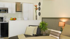 Une chambre d’hôtel compacte comprenant un coin cuisine, un lit avec deux oreillers, une table de chevet avec une lampe et une plante en pot.