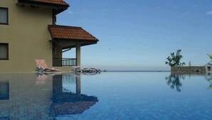 A clear blue infinity pool with lounge chairs and a building with a tiled roof. - Brand New Apartment with pool, gym and playground (Kamchia)