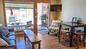 A cozy living area with a wooden bench, a sofa, a small table, and a chair. A sliding glass door leads to an outdoor patio with a barbecue grill and outdoor furniture. - Starboard Cabana - Unit 2 (Chelan)