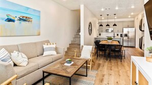 Una sala moderna con sofá, mesa de centro y comedor. Se ve una cocina con electrodomésticos y una escalera al fondo.
