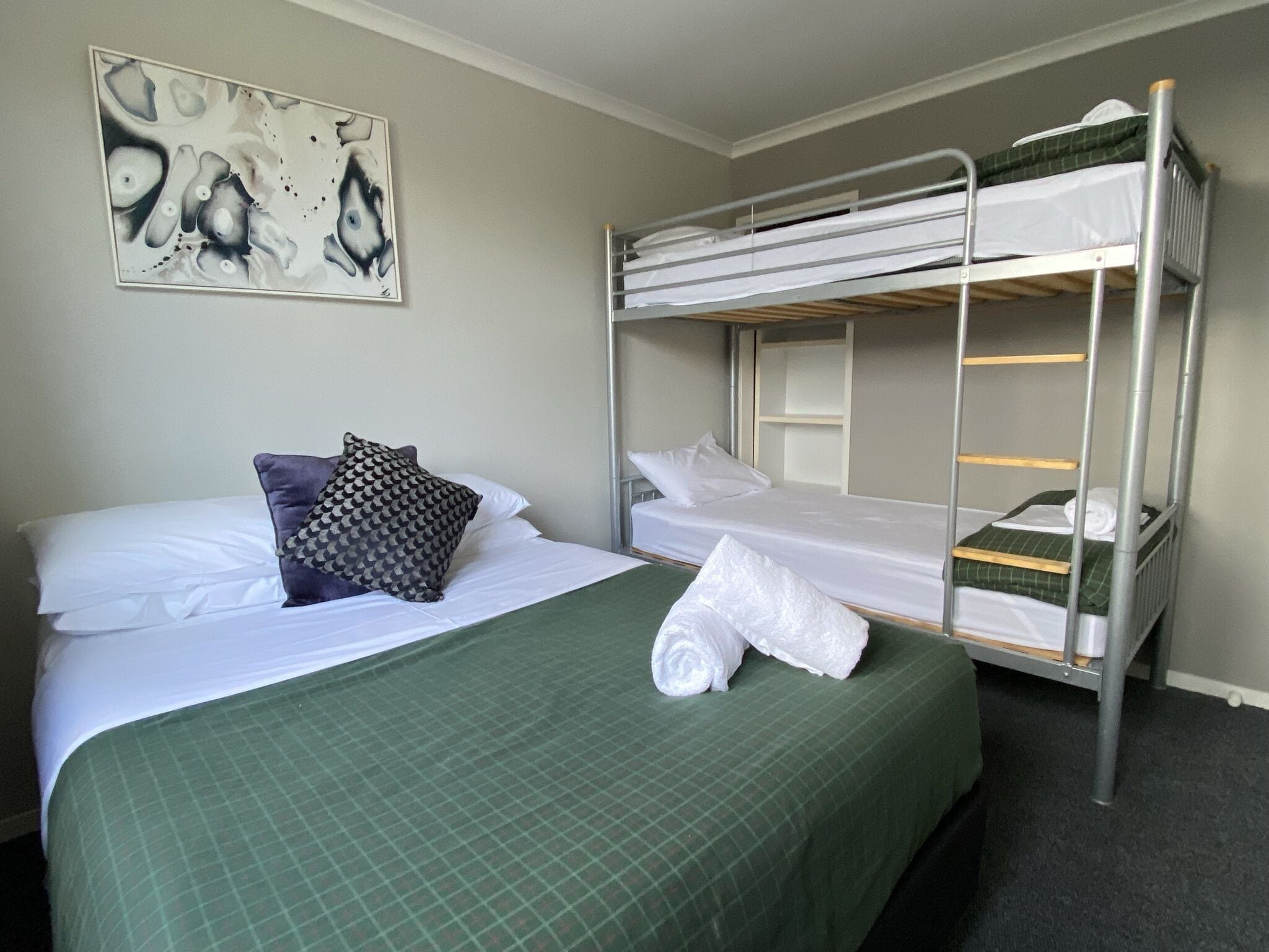 A hotel room with a bunk bed, a single bed, a green checkered bedspread, white linens, a patterned pillow, and a painting on the wall.