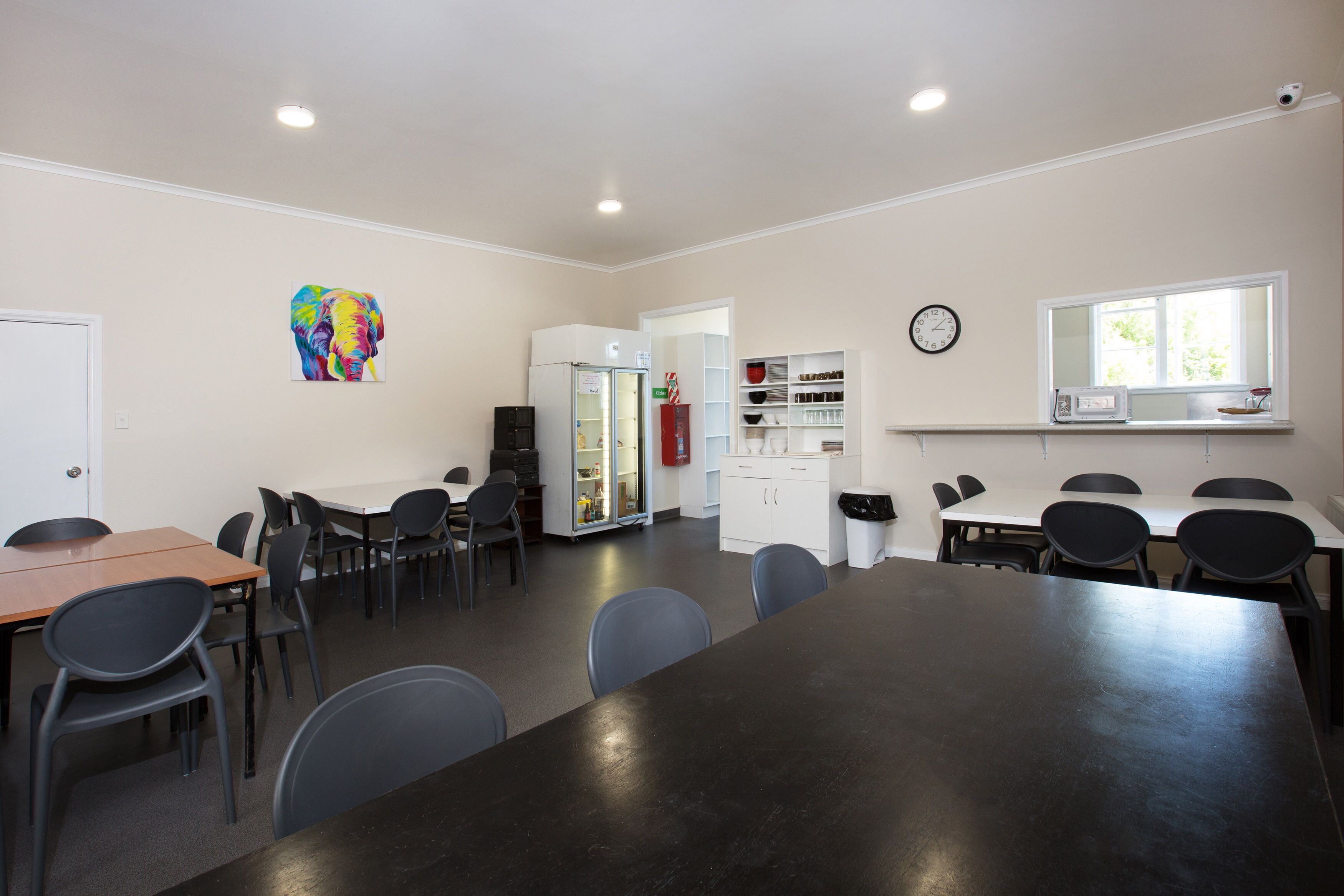Un salón de conferencias con una larga mesa, múltiples sillas, un refrigerador, un reloj y una ventana con luz natural.