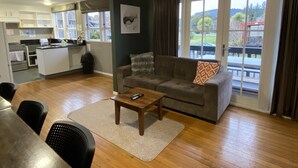 Una sala de estar con un sofá, una mesa de centro y un comedor con sillas y una mesa. Se observa una cocina al fondo, con armarios y una ventana.