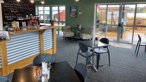 Una cafetería con un menú en pizarra que anuncia bebidas calientes y batidos, una barra con diversas opciones de bebidas, y una zona de estar con mesas y sillas.