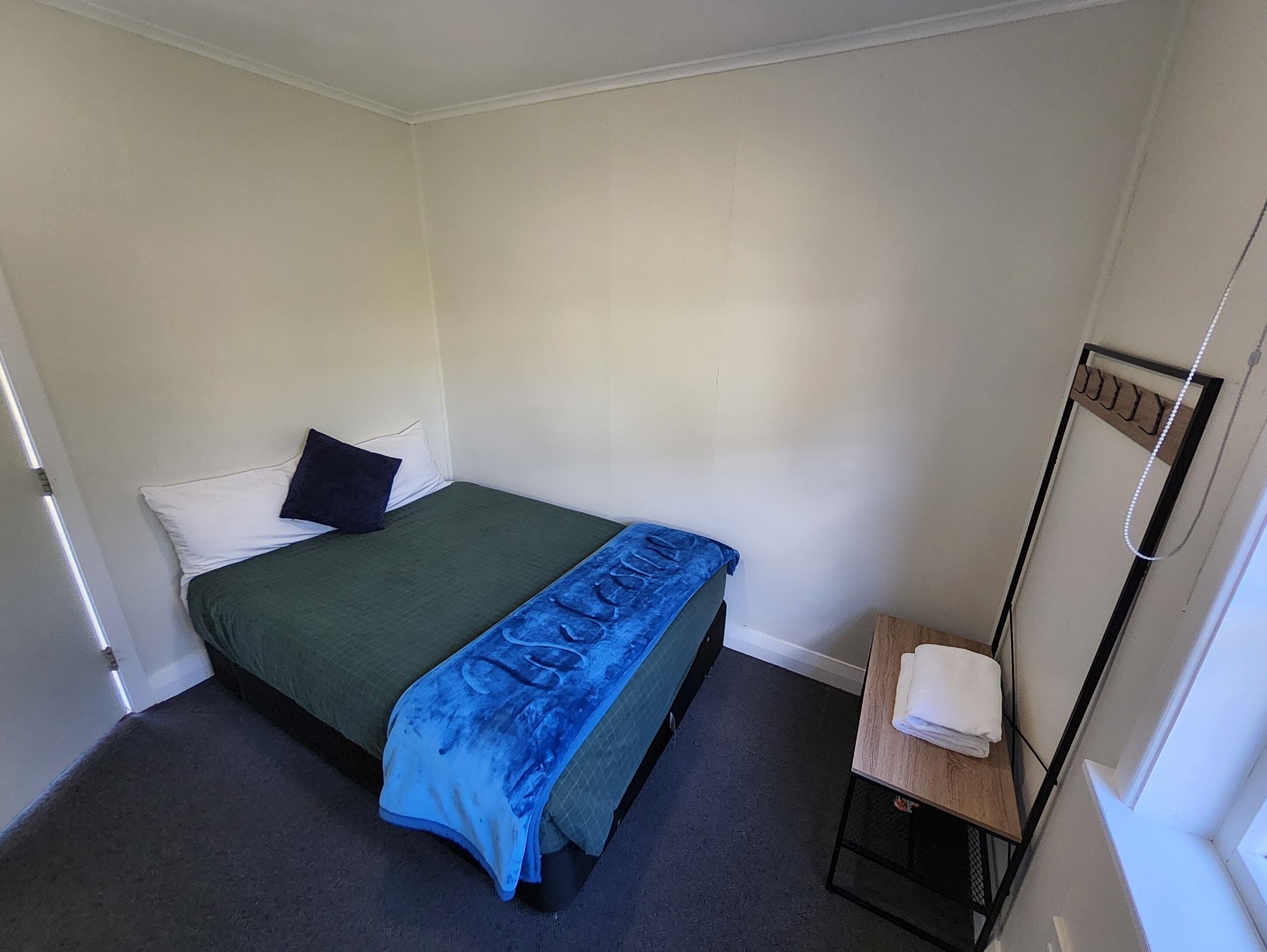 Ein kleines, sauberes Zimmer mit einem Einzelbett, einem kleinen Tisch mit Handtuch und einem Fenster mit Vorhang.