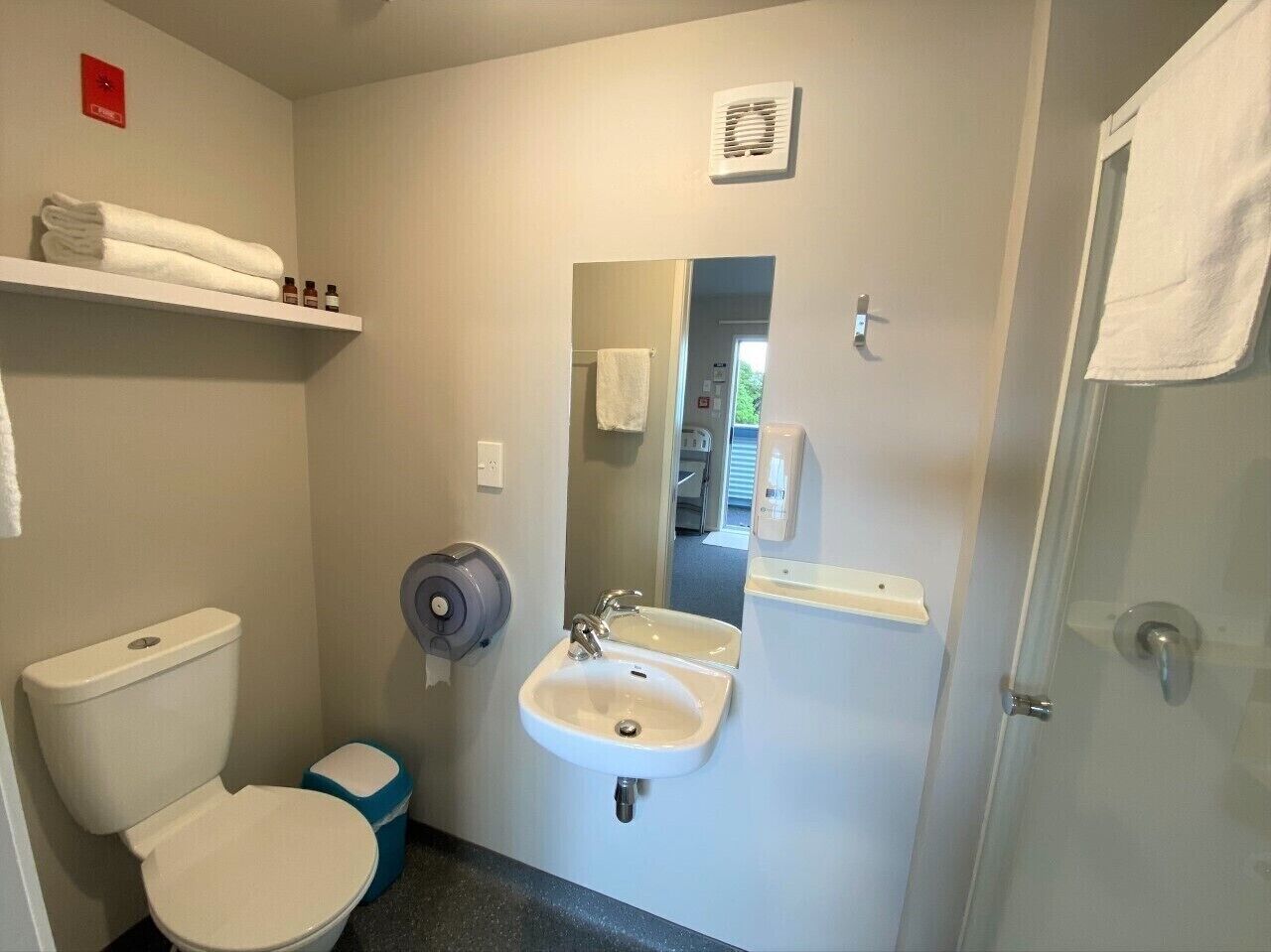 A small bathroom with a toilet, sink, and shower. There are towels on a shelf and a mirror above the sink.