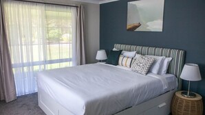 A neatly made bed with white linens, a striped headboard, and multiple pillows. There is a large window with sheer curtains, a bedside lamp, and a woven side table. - Clarence Homestead - Swim & Relax in the Mountains (Clarence)