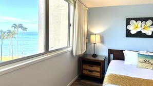 A hotel room with a large window offering a view of the ocean and palm trees, a bed with a pillow labeled "Aloha", a nightstand with a lamp, and a wall-mounted picture of a flower. - Spacious 2bedroom/2bath Beach Front Condo (Lahaina)