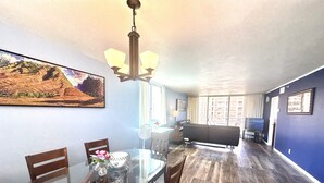 A dining area with a glass table and wooden chairs, a chandelier, and a painting on the wall. - Spacious 2bedroom/2bath Beach Front Condo (Lahaina)