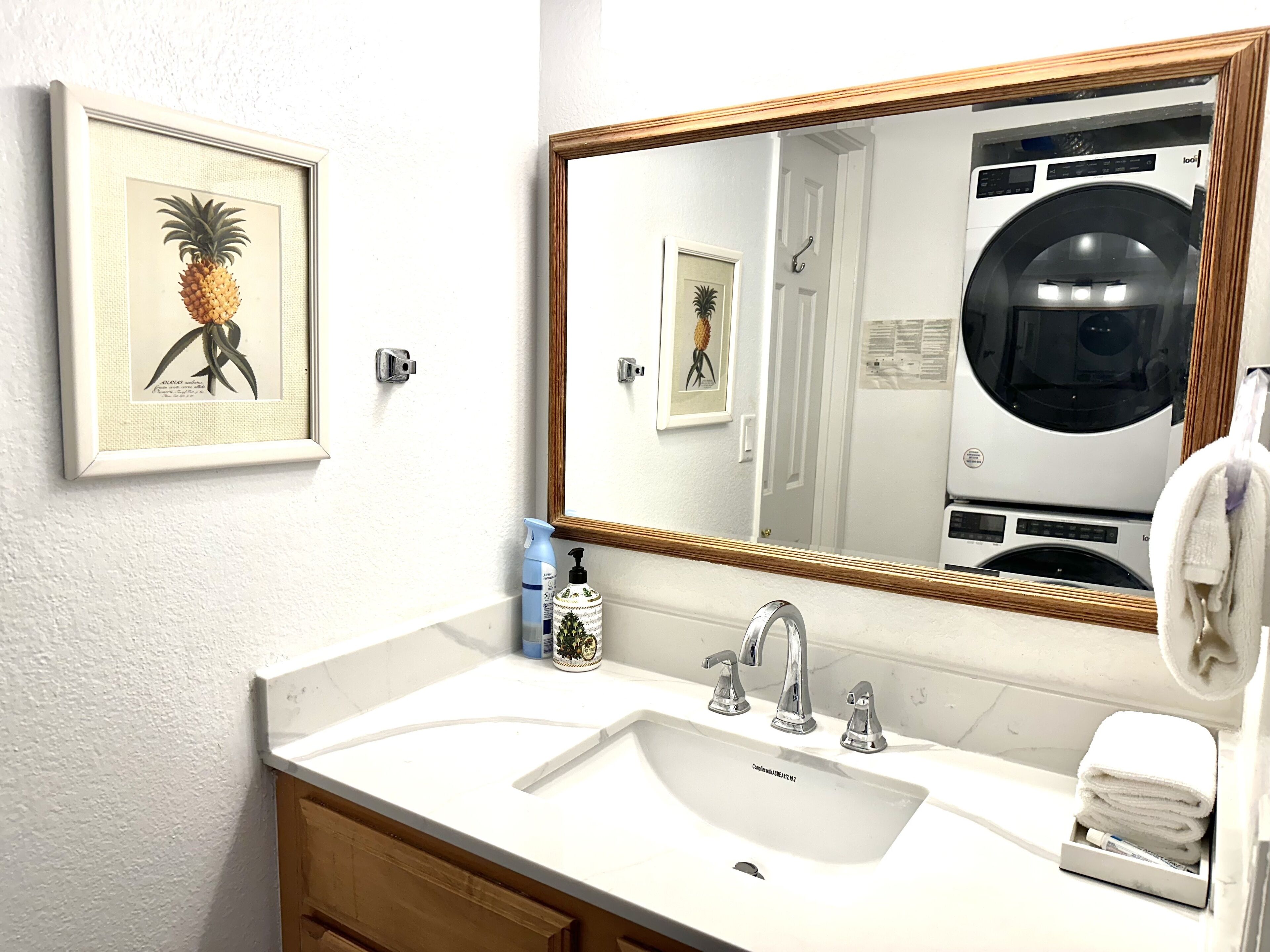 A bathroom with a white sink, two faucets, a mirror with a wooden frame, and a framed pineapple artwork on the wall.