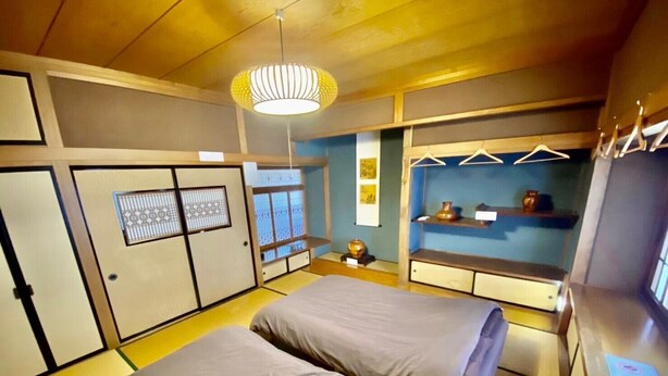 Una camera in stile tradizionale giapponese con pavimento in tatami, due letti con futon, una lampada a sospensione e uno scaffale con oggetti.