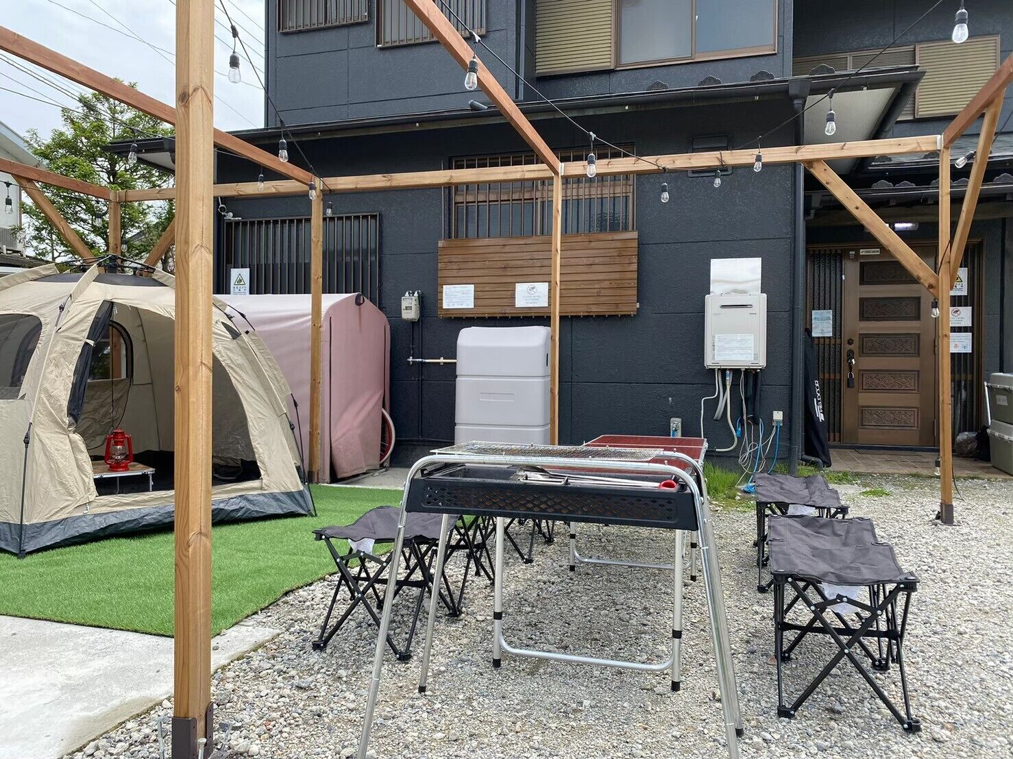 A camping setup with a tent, portable toilet, and camping chairs in front of a building with a black exterior and wooden beams.