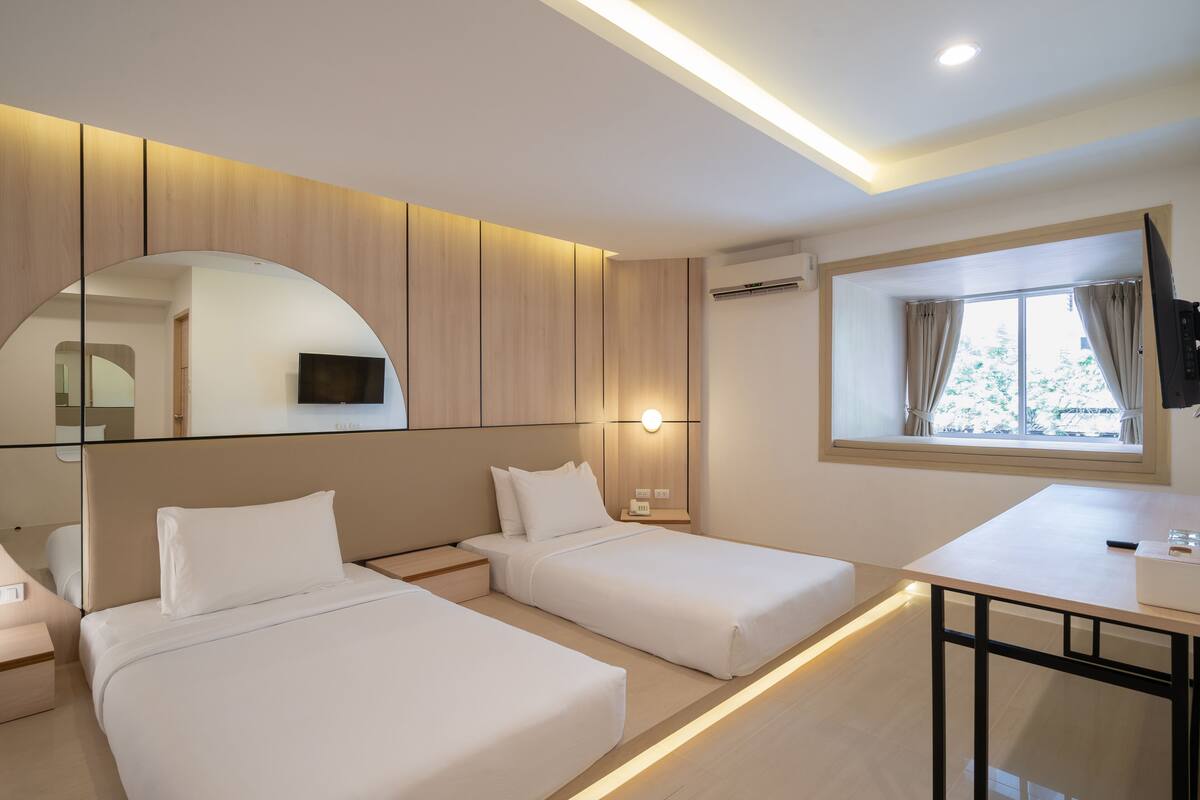 A modern hotel room with two single beds, a desk, a TV, and a large window with curtains.