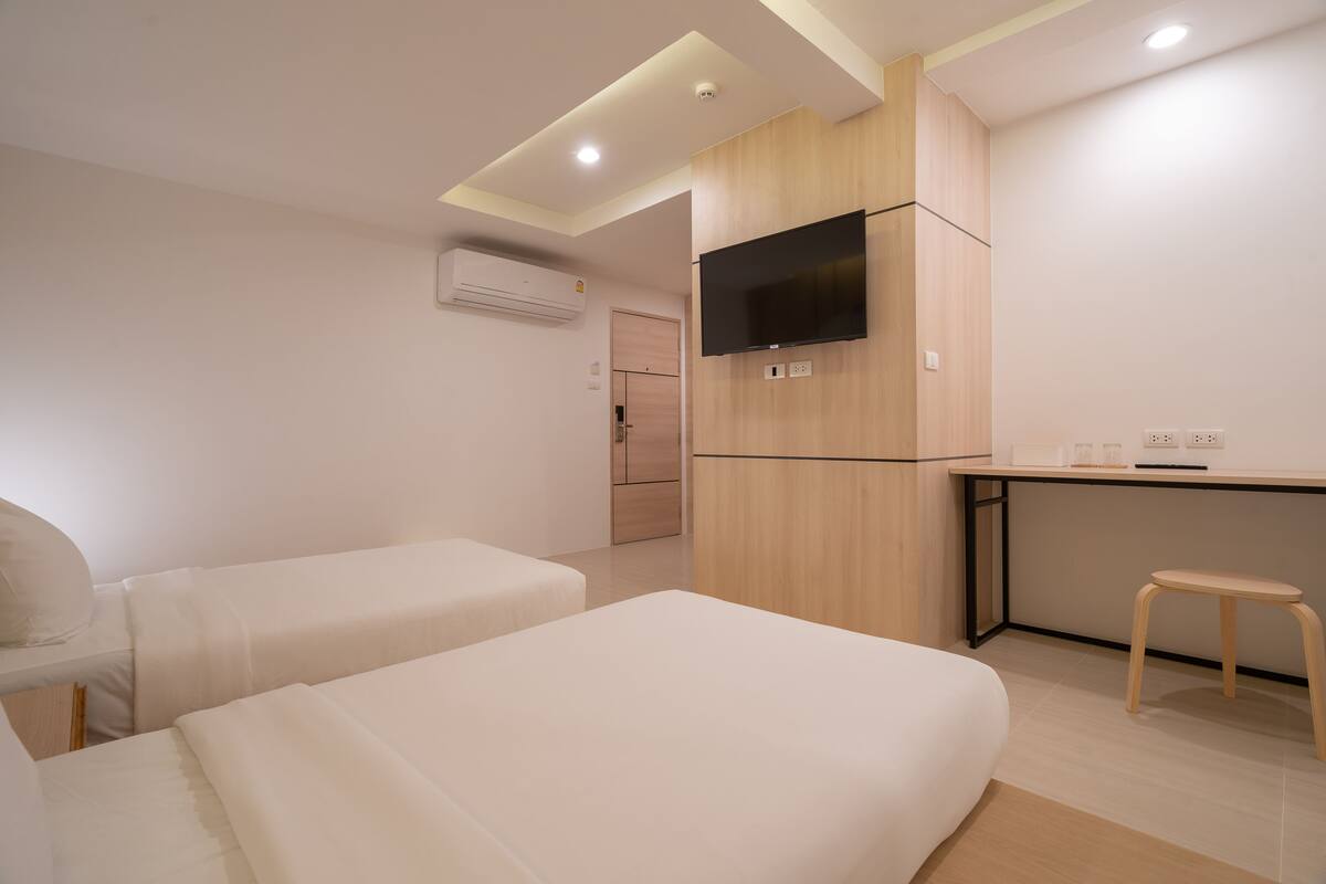 A modern hotel room with two beds, a desk, a TV, and a small stool.
