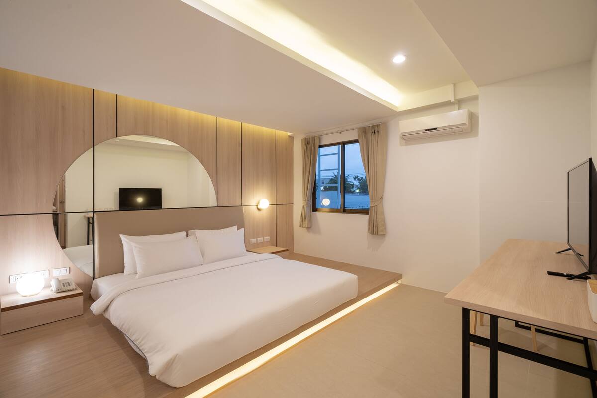 A modern hotel room with a large bed, bedside table with a lamp, a desk with a chair, a TV, and a window with curtains.