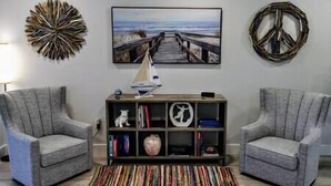 Un salon avec deux fauteuils gris, une table basse en bois, une bibliothèque et un tableau représentant une jetée.