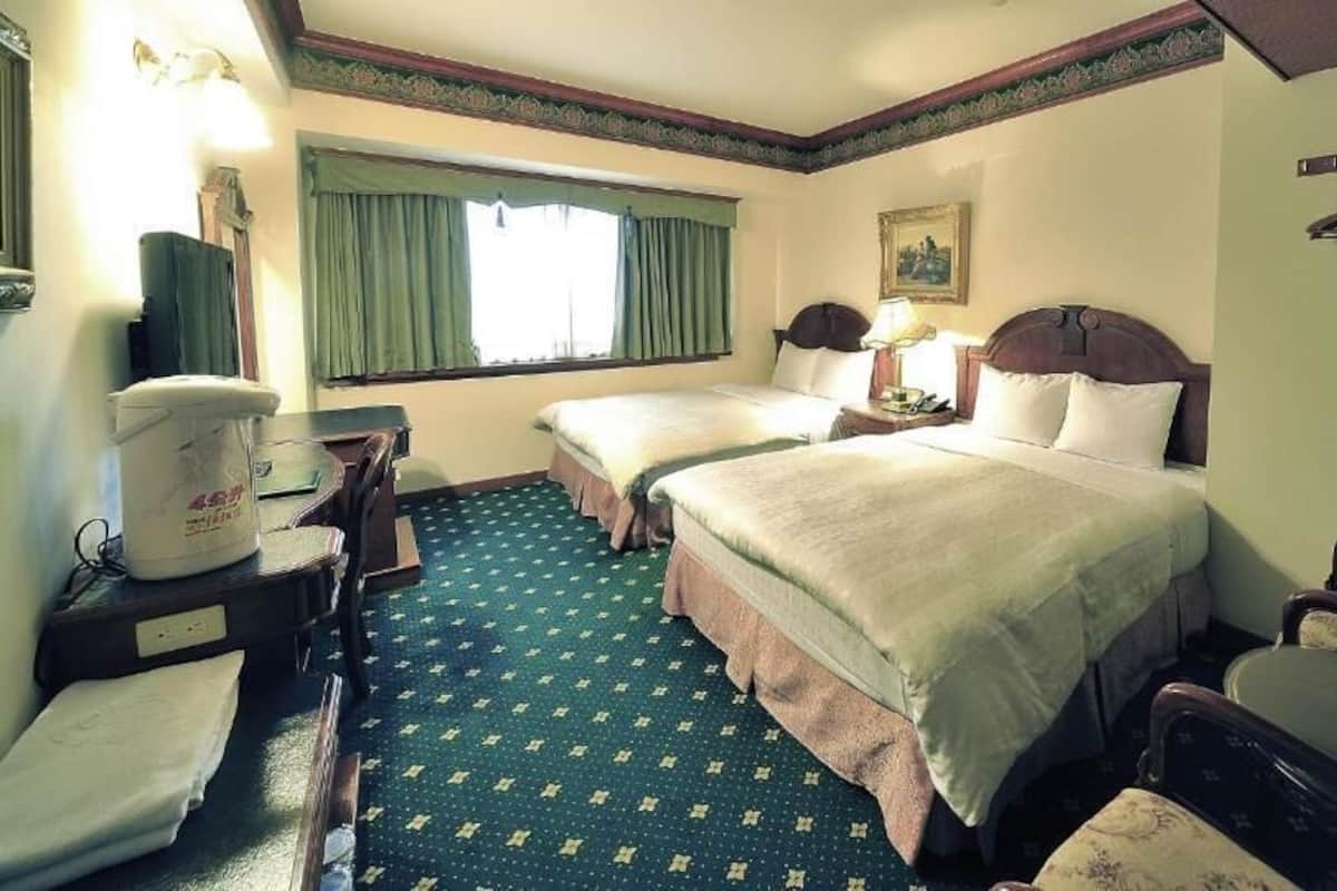 Ein Hotelzimmer mit zwei Betten, einem Schreibtisch, einem Stuhl, einem kleinen Tisch, einem Kleiderhaken und einem Fenster mit Vorhängen.