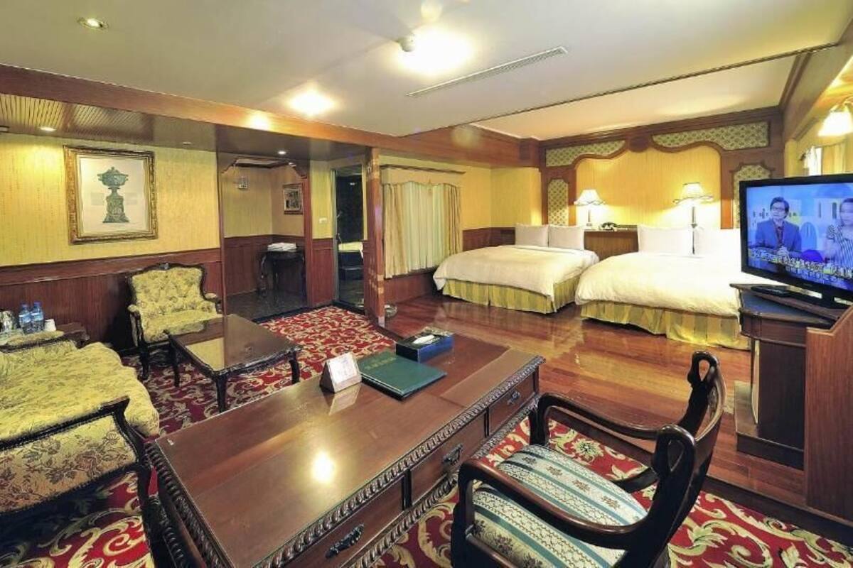 Ein Hotelzimmer mit Holzschreibtisch, zwei Stühlen, einem Bett, einem Fernseher und einem kleinen Tisch mit einem Buch und Wasserflaschen.