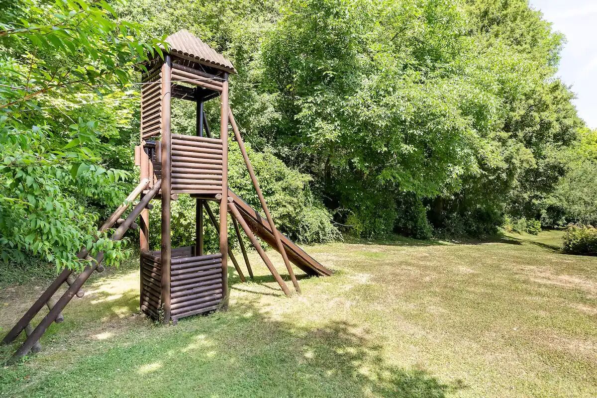 Une structure de jeu en bois, dotée d’un toboggan et d’éléments d’escalade, installée dans un espace herbeux entouré d’arbres.