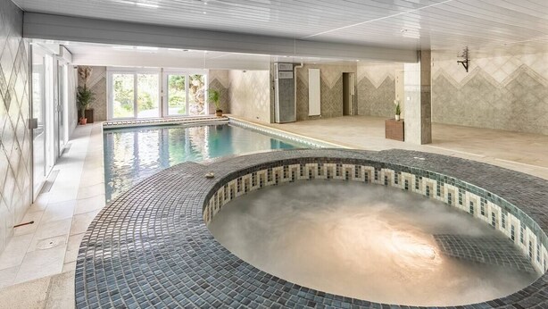 Ein großer Innenpool mit einem gefliesten Whirlpoolbereich, umgeben von einer gefliesten Liegefläche und einer Glasumrandung.