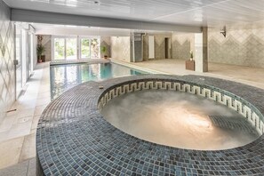 Een groot binnenzwembad met een betegeld hottubgedeelte, omringd door een betegeld zonnedek en een glazen overkapping.