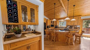 A spacious kitchen with wooden cabinets, a central island with stools, and a large window providing natural light. - Kubo Del Sol - Log Cabin, Hot Tub, Game Room, Pool Table, Ping Pong (Big Bear Lake)