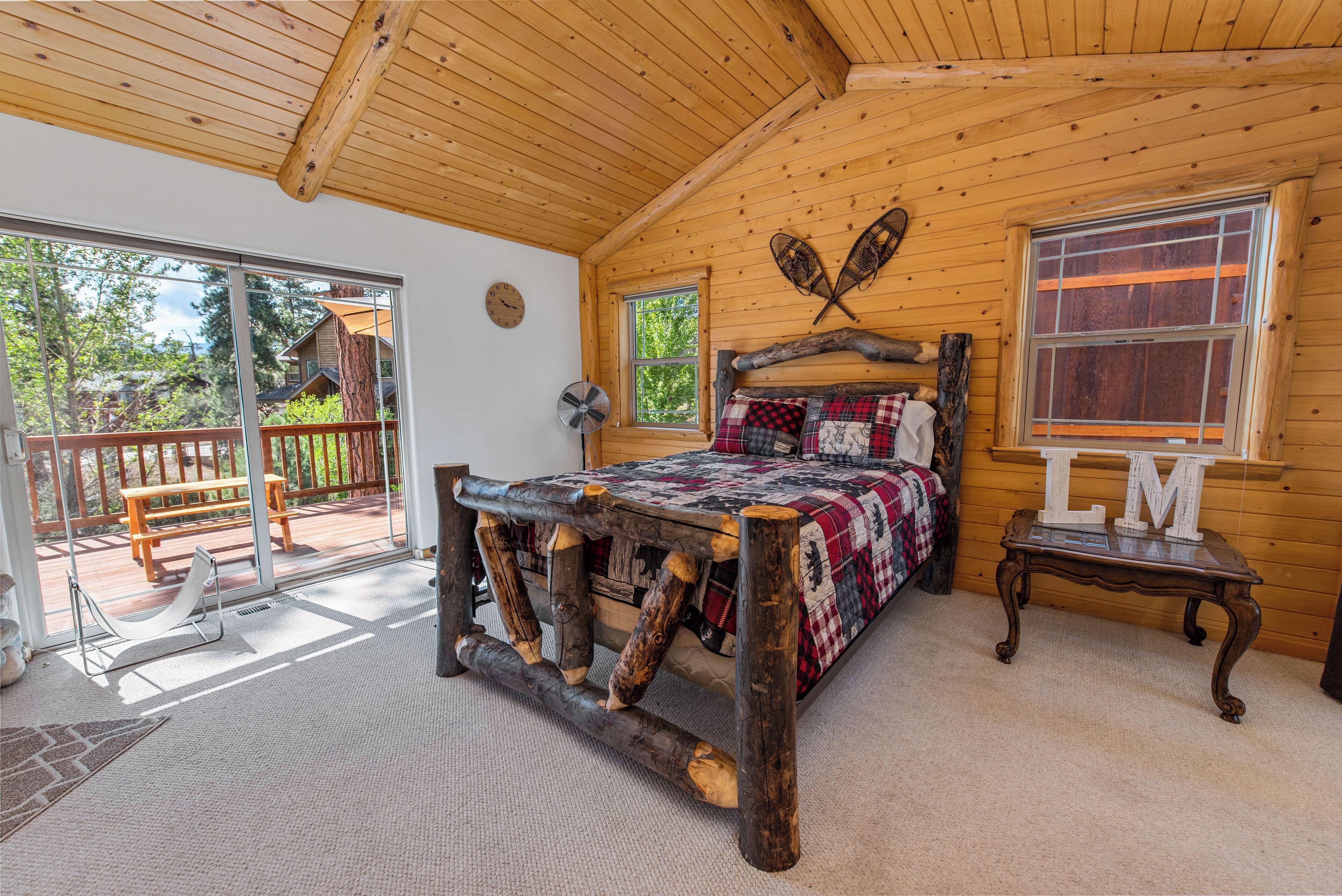 Une chambre à coucher dans une cabane en bois, avec un lit, une tête de lit en bois, une fenêtre ornée des lettres « LM » et une vue sur l’extérieur.