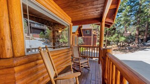 A wooden cabin with a porch, rocking chairs, and a stone wall visible through the window. - Kubo Del Sol - Log Cabin, Hot Tub, Game Room, Pool Table, Ping Pong (Big Bear Lake)