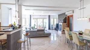 Eine moderne Küche mit einer zentralen Kochinsel, Essbereich mit Glastisch und Stühlen sowie Wohnbereich mit Sofa und Fernseher.