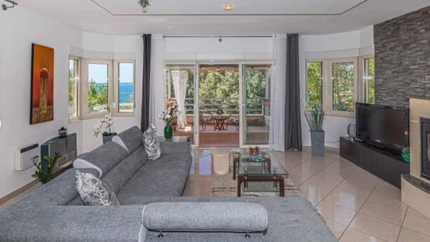 Ein modernes Wohnzimmer mit einem grauen Ecksofa, einem Glastisch und einem Flachbildfernseher. Ein Balkon bietet Blick ins Freie.