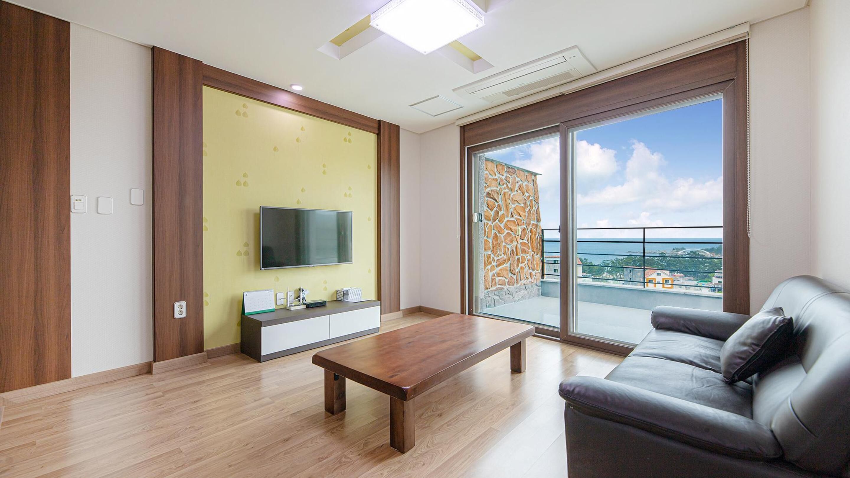 A modern living room with a wooden coffee table, a grey sofa, a TV mounted on a yellow wall, and a balcony with a stone wall.