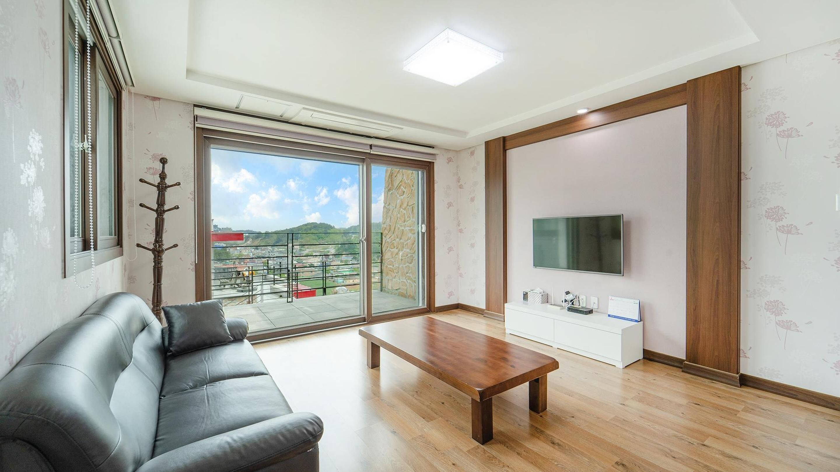 A living room with a sofa, a coffee table, a TV, and a sliding glass door leading to a balcony with a view of a cityscape.
