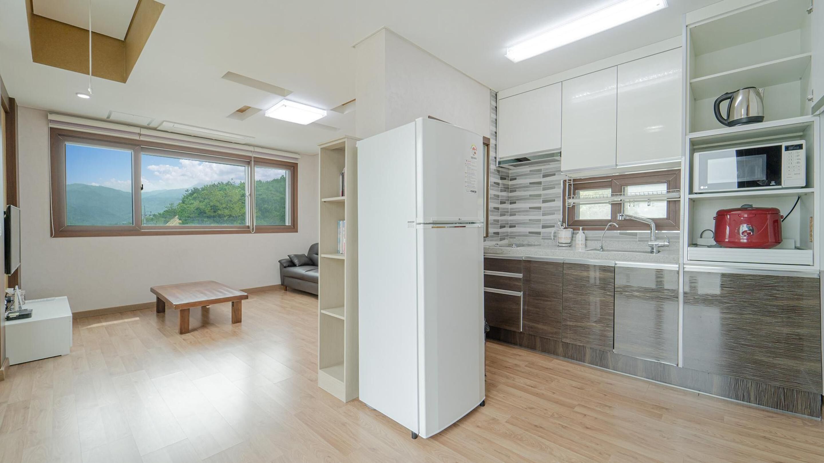 A modern kitchen with a built-in fridge, microwave, and red slow cooker. There's a large window with a view of greenery and mountains.