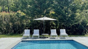 Una piscina con cuatro sillas de descanso blancas y una mesa bajo un paraguas grande, rodeada de una valla verde exuberante y árboles.