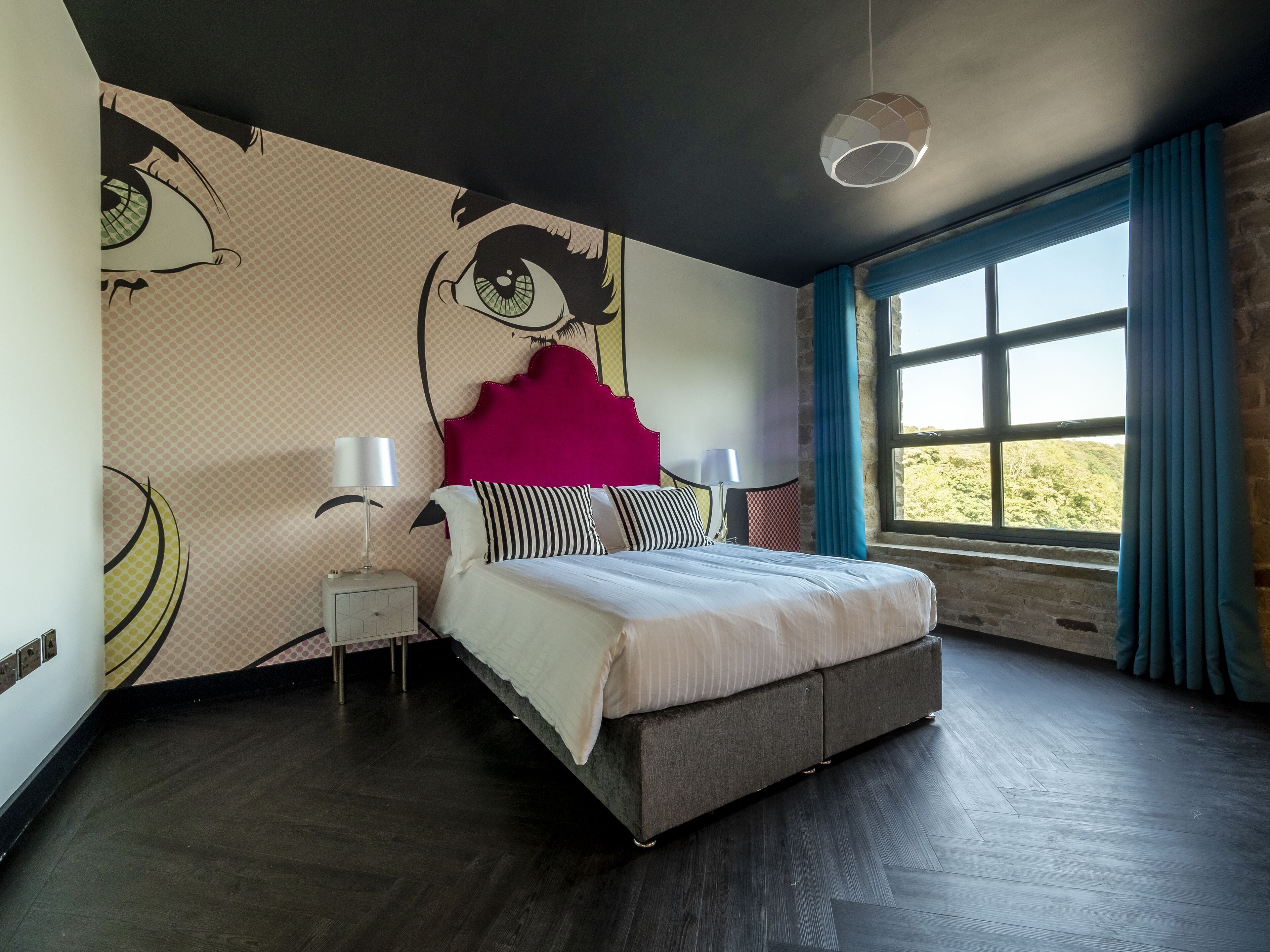 A bedroom with a large bed, a headboard with a red accent, a wall mural, a window with blue curtains, and a modern light fixture.