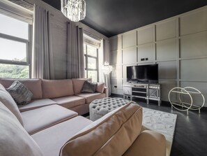 A modern living room with a large sectional sofa, a tufted ottoman, a flat-screen TV on a cabinet, and a chandelier.