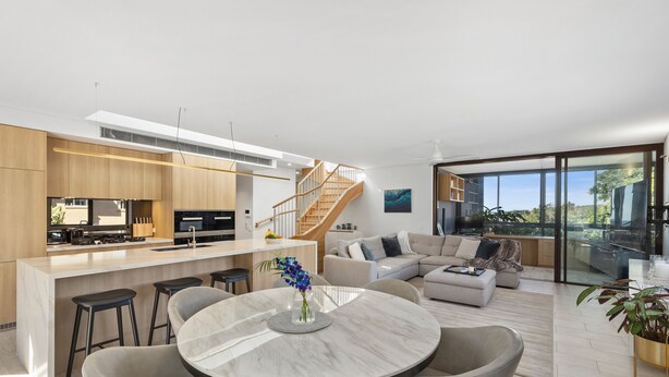 Eine moderne Küche mit einer zentralen Kochinsel, ein Essbereich mit Marmortisch, ein Wohnzimmer mit Sofa und Fernseher sowie eine Treppe, die in eine obere Ebene führt.